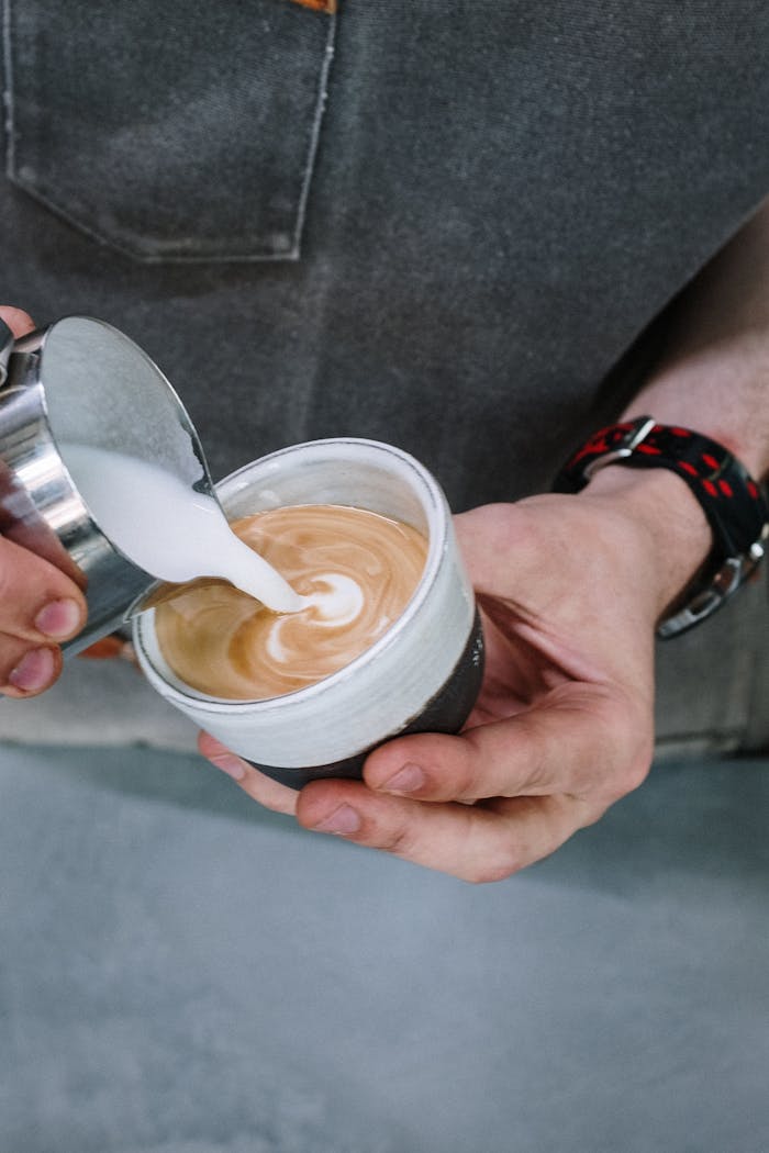 about-img Close-up of a barista pouring milk to create latte art in a cozy café setting.